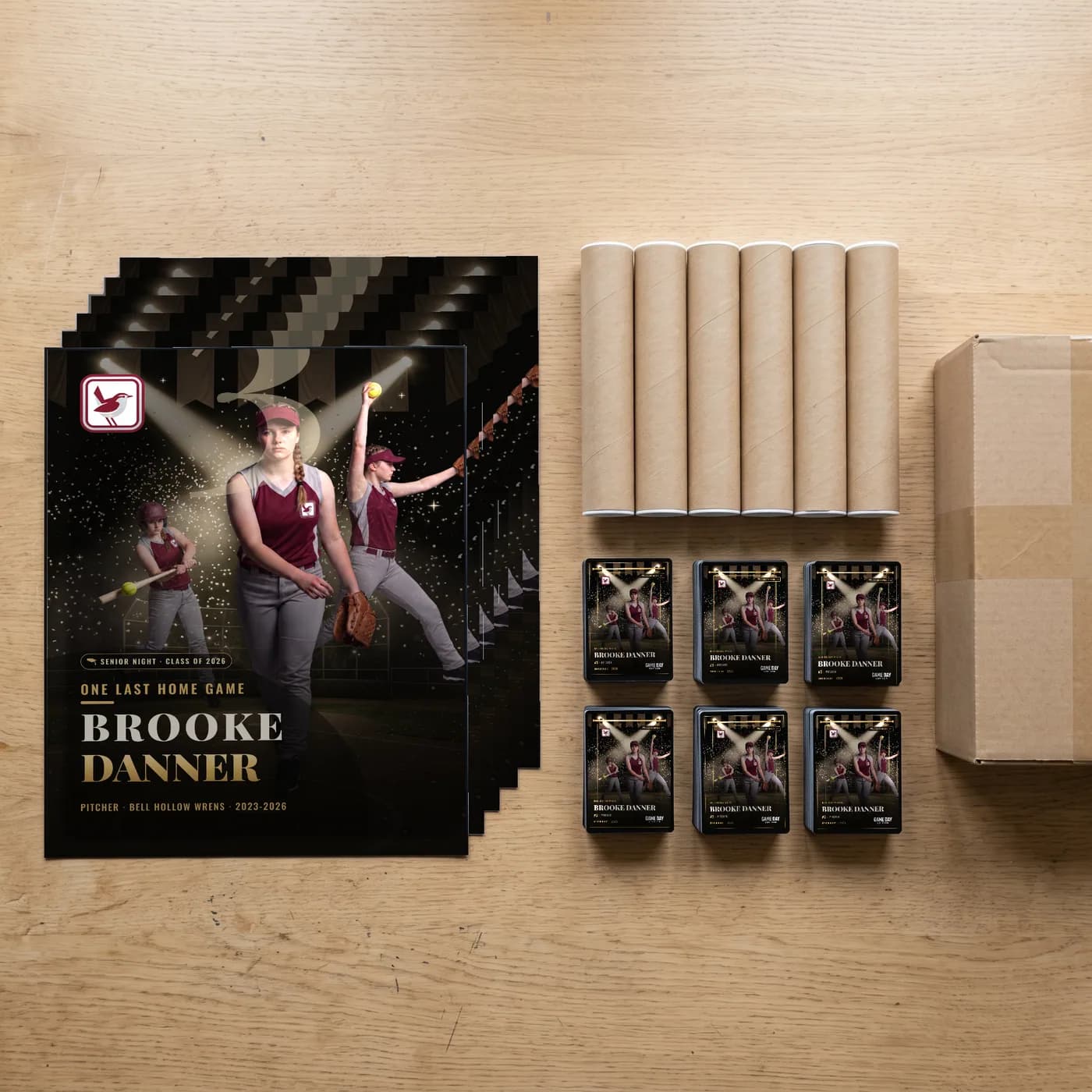 A team order staged on a table: six custom softball posters, six shipping tubes, six stacks of trading cards and the box they ship in — Senior Night finish — example artwork, fictional athlete; photo generated